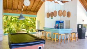 Una mesa de billar con un reloj encima, rodeada de taburetes de madera y una zona de bar con refrigerador de Pepsi.