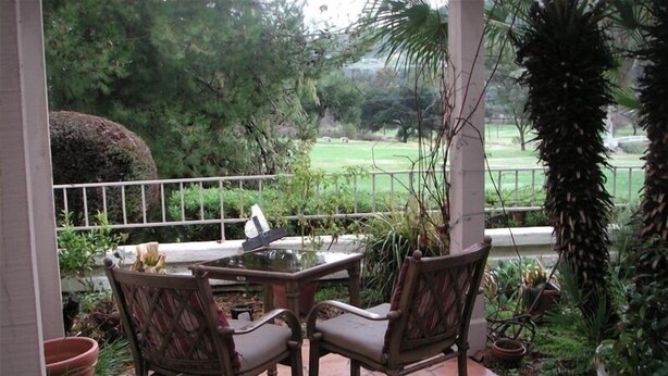 An outdoor patio with a glass-top table and two chairs, surrounded by potted plants and a view of a golf course. - Cool Summers + Warm Winters on 18 Hole Golf Course (Escondido)