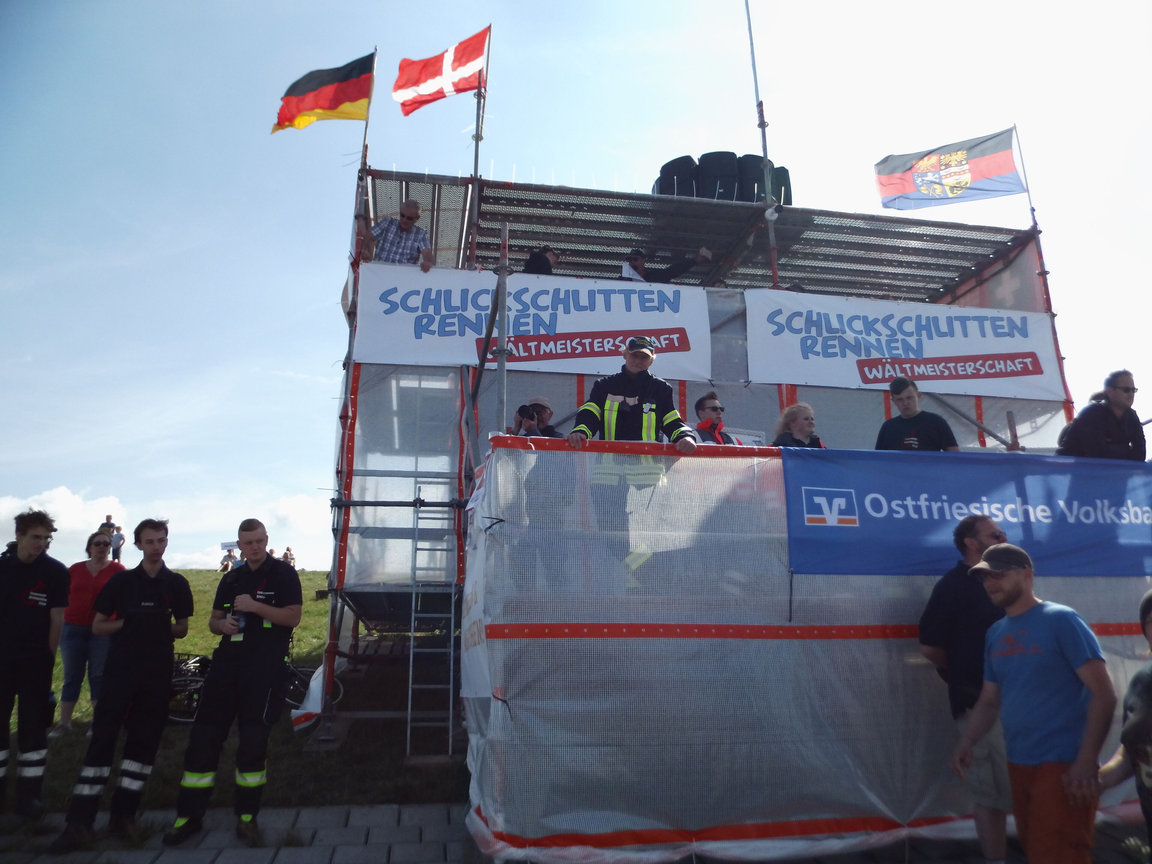A group of people standing in front of a structure with banners displaying "Schlachtschlitten Rennen" and "Ostfriesische Volkswagen".
