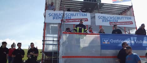 A group of people standing in front of a structure with banners displaying "Schlachtschlitten Rennen" and "Ostfriesische Volkswagen".