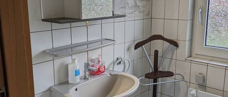 Une salle de bain moderne avec un lavabo blanc, un miroir et des étagères. Il y a une fenêtre donnant sur des arbres et une porte avec une poignée.