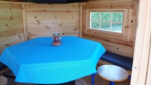A wooden cabin interior with a round table covered by a blue tablecloth, a small stool, and a window with a view of greenery. - Apartment at the old Greetsieler Tief (Eilsum)