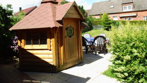 A wooden playhouse with a pitched roof and a chimney, surrounded by greenery and a paved area. - Apartment at the old Greetsieler Tief (Eilsum)