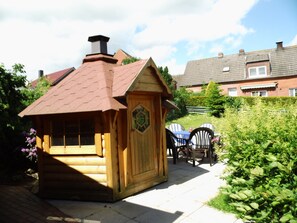 Een houten speeltuintje met een schuin dak en schoorsteen, omringd door groen en een geplaveid terras.