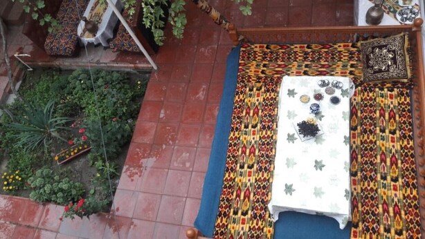 Une pièce avec un tapis à motifs, un lit recouvert d’un drap blanc, un petit espace jardin avec des plantes et des fleurs, et une table sur laquelle se trouvent divers objets.