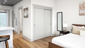 A modern hotel room with a wooden floor, a white bed, a small bedside table with a lamp, a large mirror, and a painting on the wall. - Studio King Suite with Kitchen | 202 (Asheville)
