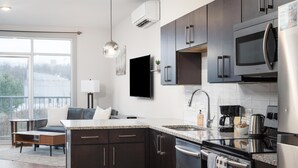 A modern kitchen with dark cabinetry, a stainless steel refrigerator, a microwave, and a dishwasher. There is a large window providing natural light and a view of the outdoors. - Studio King Suite with Kitchen | 202 (Asheville)