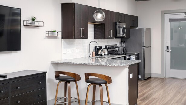 A modern kitchen with dark cabinets, a stainless steel refrigerator, a microwave, and a granite countertop. There are two wooden bar stools and a hanging pendant light. - Studio King Suite with Kitchen | 202 (Asheville)