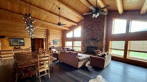 A log cabin interior with a dining area, seating, a fireplace, and large windows. - Black Tree Resort (Lake George)