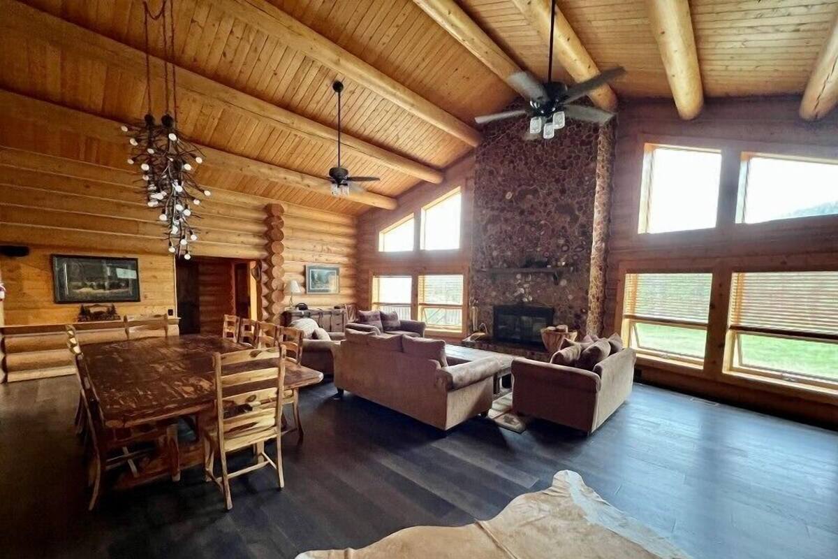 A log cabin interior with a dining area, seating, a fireplace, and large windows.