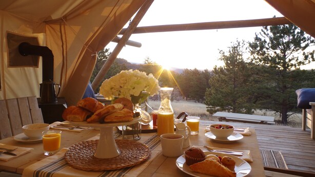 A breakfast setup in a tent with a table, breakfast items, and a view of trees and hills. - Black Tree Resort (Lake George)