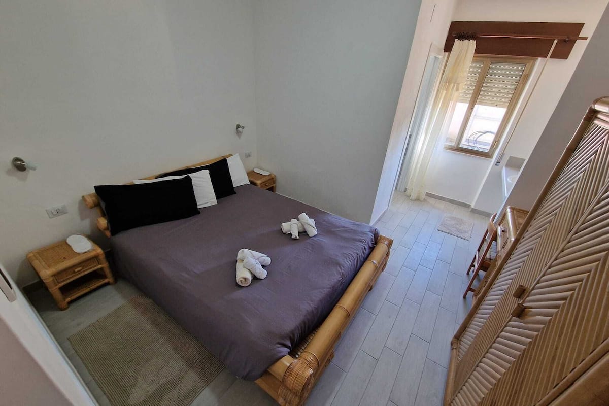 Un dormitorio con cama, almohadas, mesita de noche, armario de madera y ventana con cortinas.