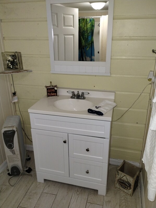 A bathroom with a white vanity, a sink, and a mirror above it. There is a small shelf with a bottle and a sign that reads "Gone Fishin'". A heater is visible on the floor to the left of the vanity.