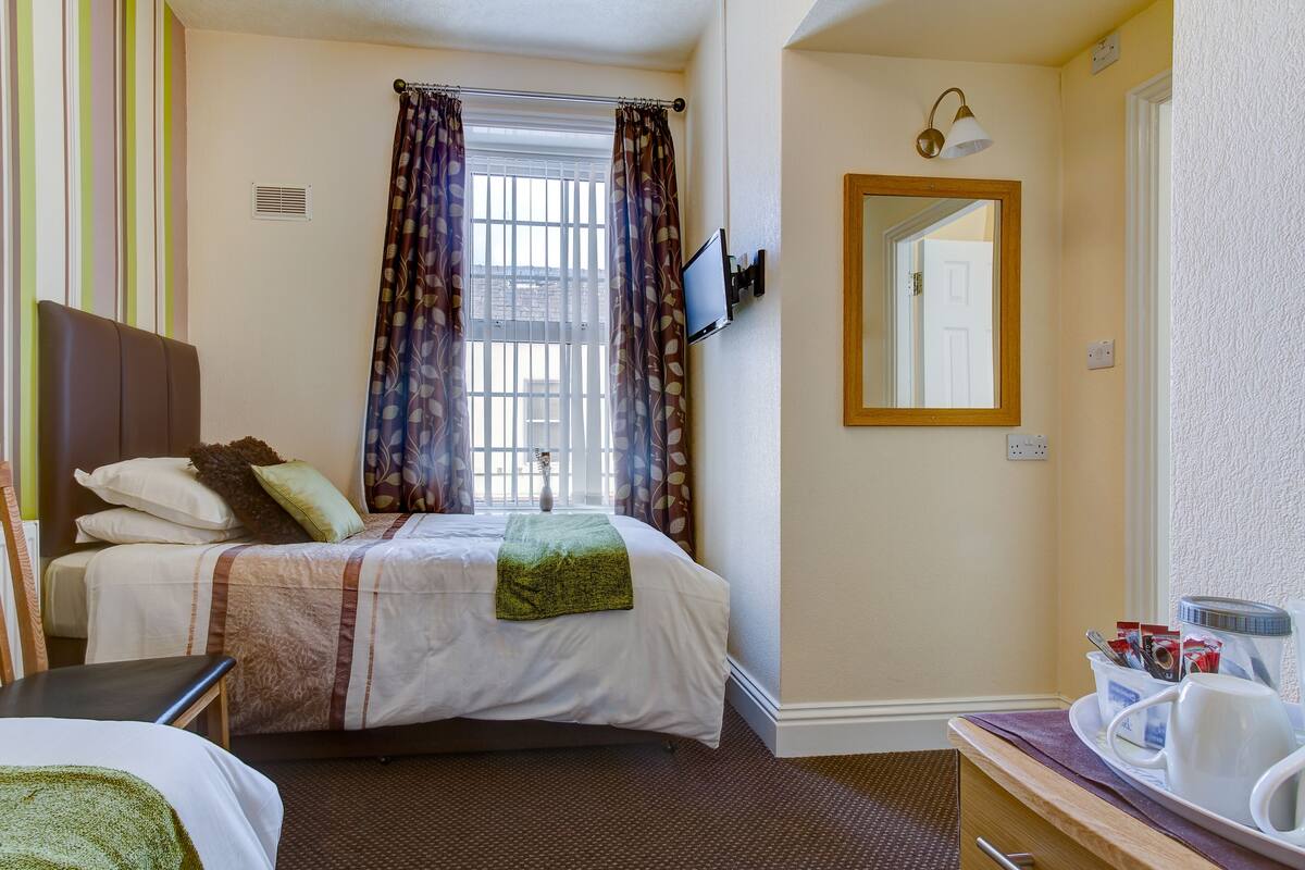 A hotel room with a bed, a TV mounted on the wall, a window with curtains, a bedside table with a tray, and a mirror.