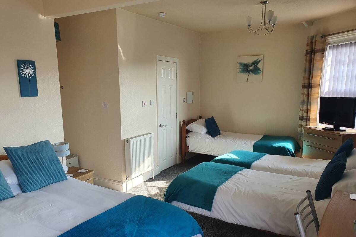 A hotel room with three single beds, a desk with a chair, a television, and a window with curtains.