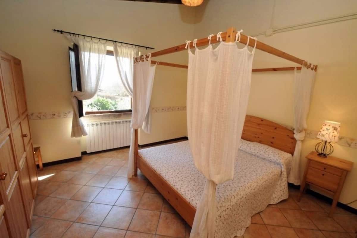 A bedroom with a four-poster bed, a wooden nightstand, a radiator, and a window with curtains.