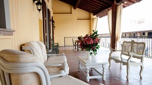 Un área de estar al aire libre, cubierta, con muebles blancos, una mesa con un florero y vistas a edificios al fondo.
