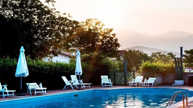 Una piscina con sillas de descanso blancas y sombrillas, rodeada de vegetación y una valla.