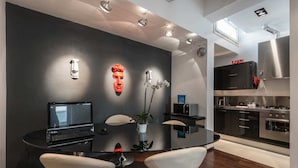 Una cuina moderna amb electrodomèstics d'acer inoxidable, zona de menjador amb taula de vidre i cadires blanques, i paret fosca amb elements decoratius.
