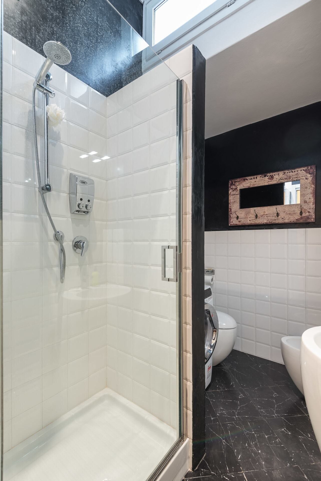 Un baño moderno con ducha de vidrio, paredes de azulejos blancos, suelo de mármol negro y un inodoro blanco.