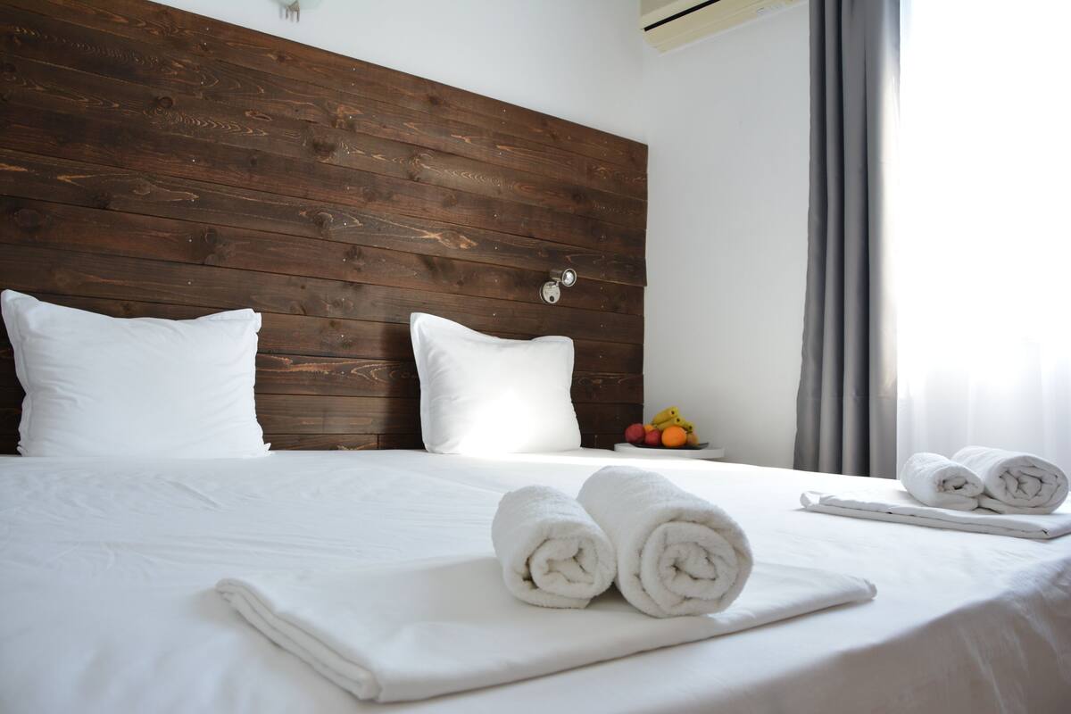A neatly made bed with white linens and towels, a wooden headboard, and a fruit bowl on the nightstand.