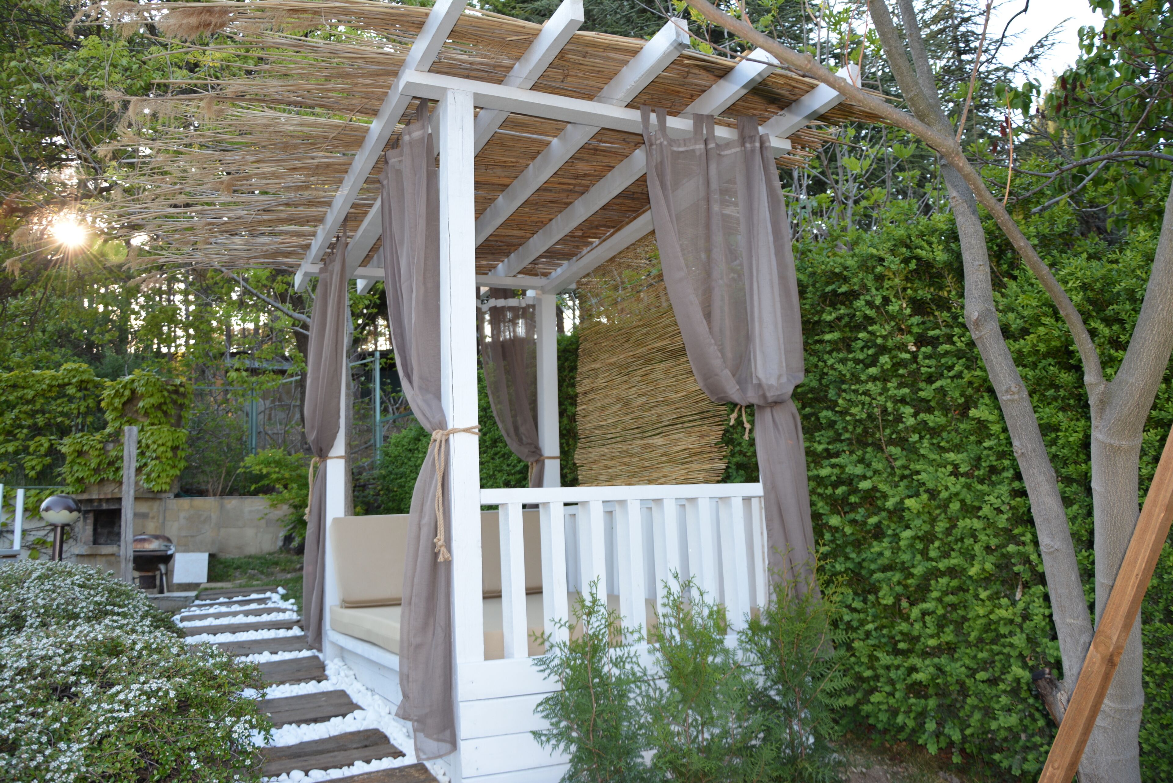 Una pergola in legno con ringhiere bianche e cuscini color beige, immersa nel verde.