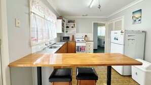 A compact kitchen with a wooden table, two stools, a microwave, a stove, and a refrigerator. - The Secret Waypoint - Kalbarri, WA (Kalbarri)