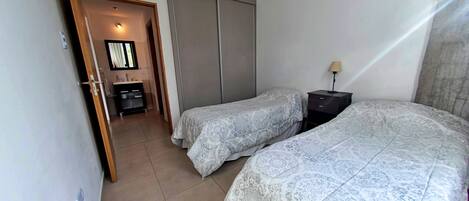 Un dormitorio con dos camas, un armario, una mesita de noche con lámpara y un baño visible a través de una puerta abierta.