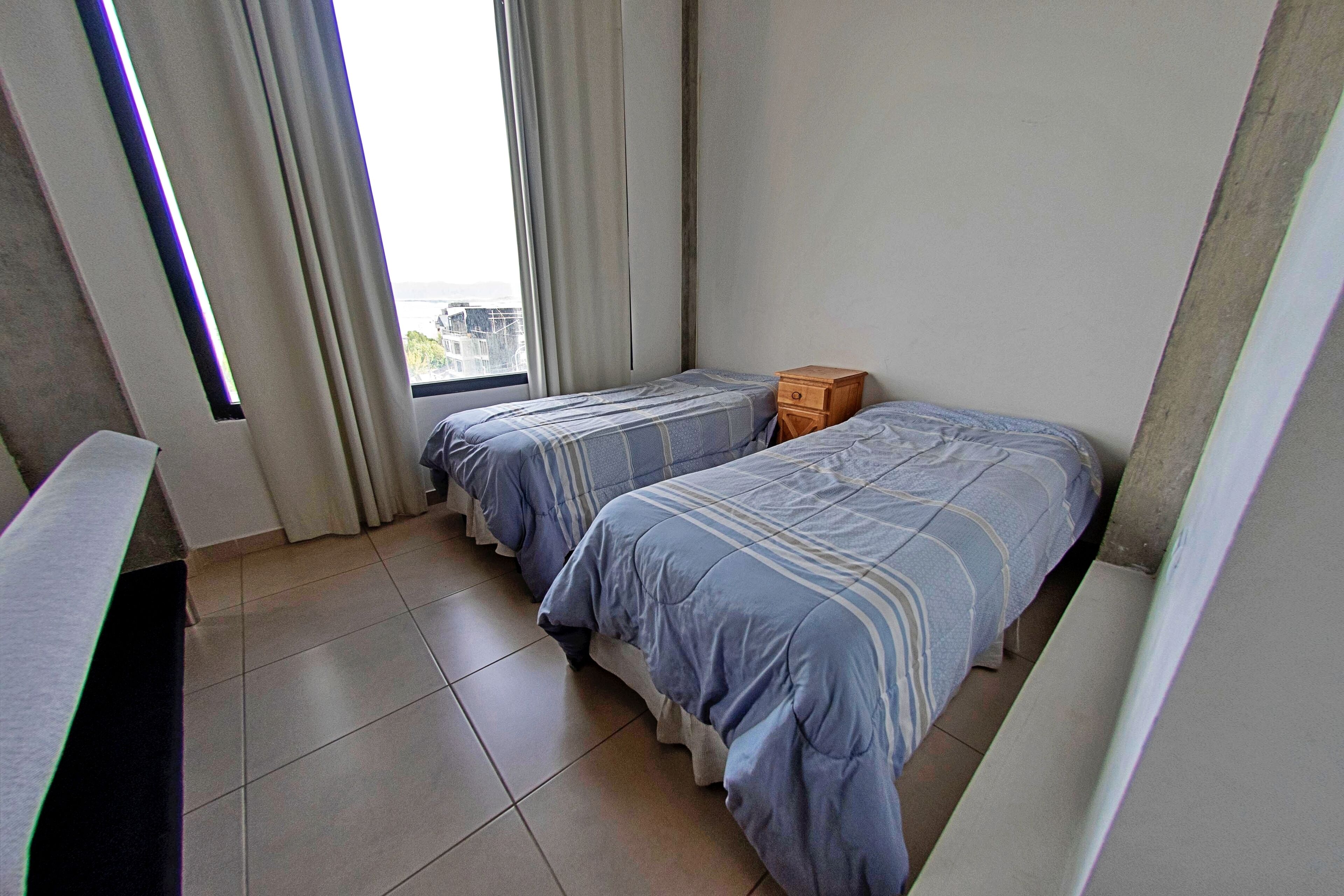 A small, clean room with two single beds, a wooden nightstand, and a window with curtains.