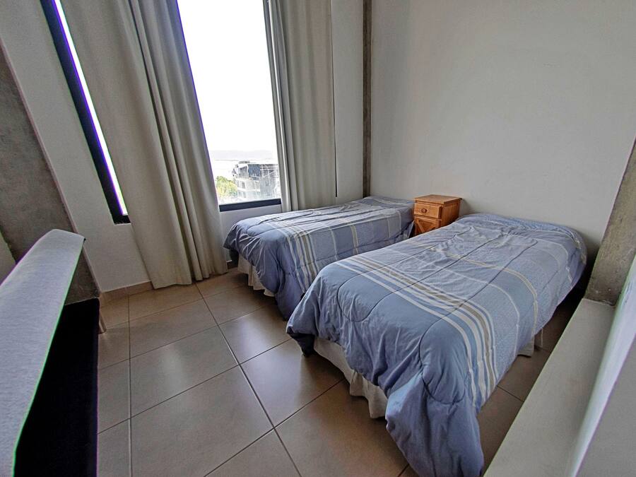 A small, clean room with two single beds, a wooden nightstand, and a window with curtains.