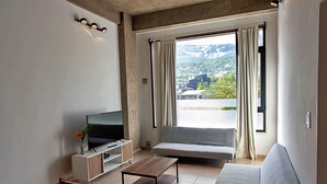 Een moderne woonkamer met een flatscreen televisie, een houten salontafel, een grijze bank en een raam met gordijnen.