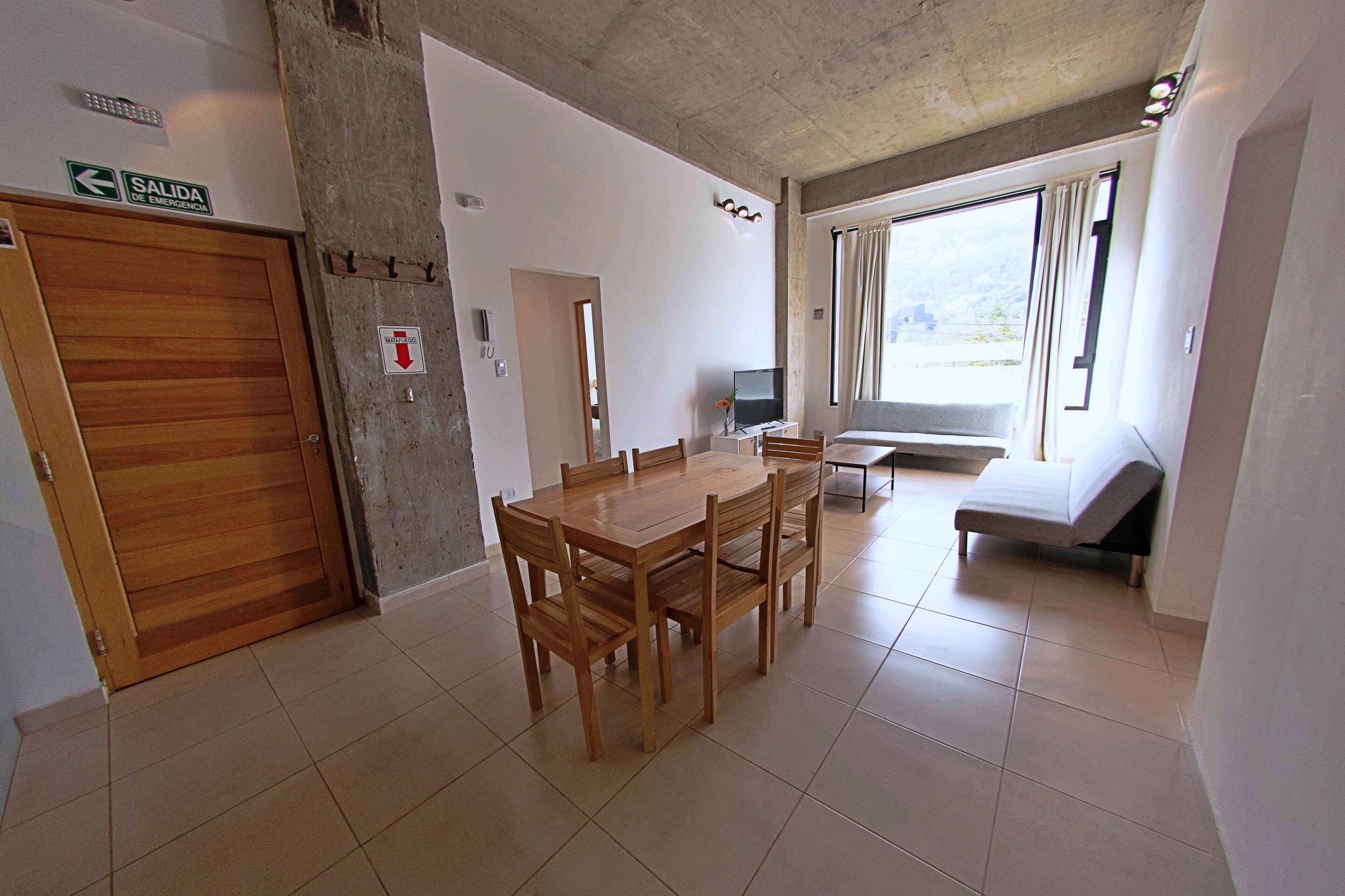 Ein Zimmer mit einem Holztisch, Stühlen, einem Fernseher, einem Sofa und einem Fenster mit Vorhängen.