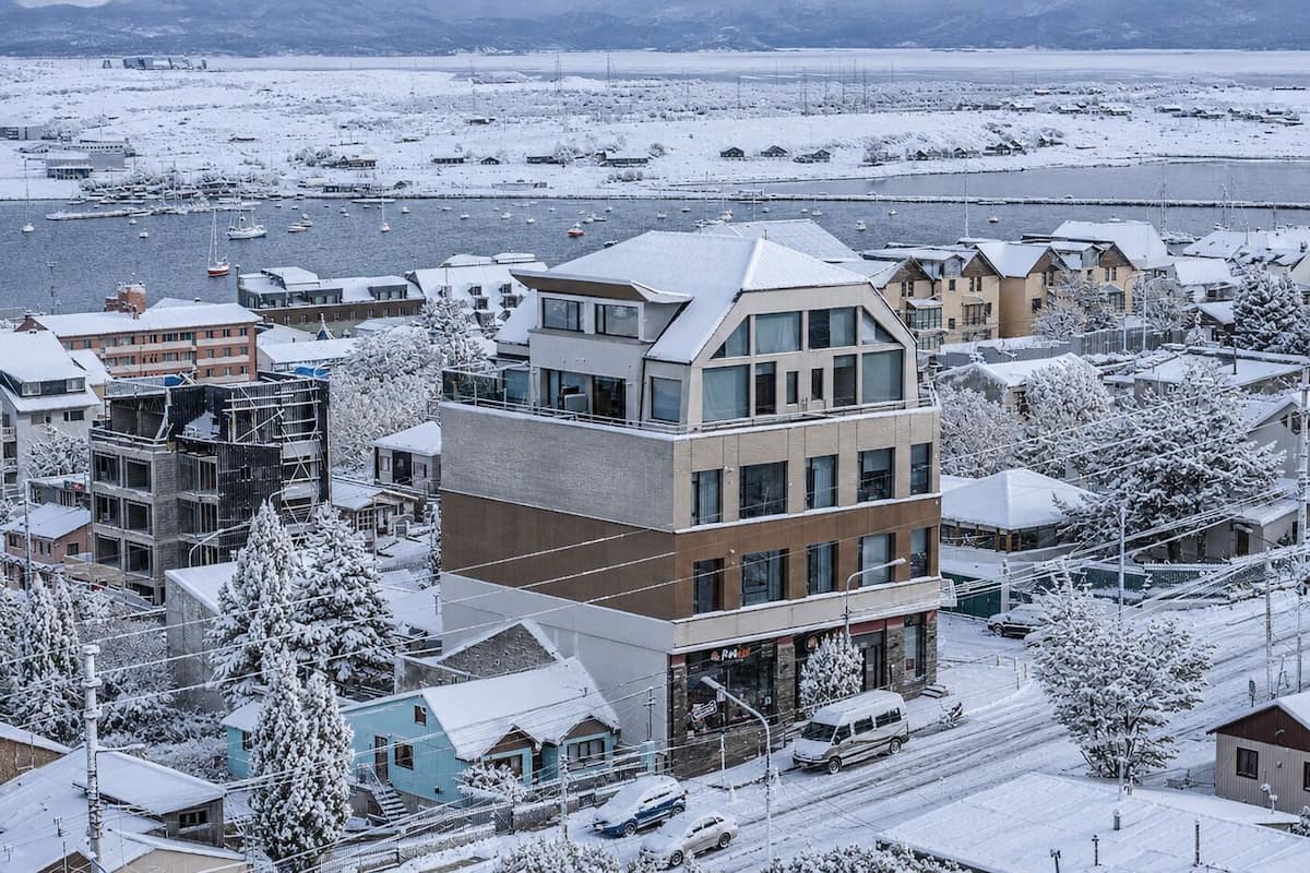 Un edificio de varios pisos con un diseño arquitectónico moderno, situado en un pueblo nevado con un río al fondo.