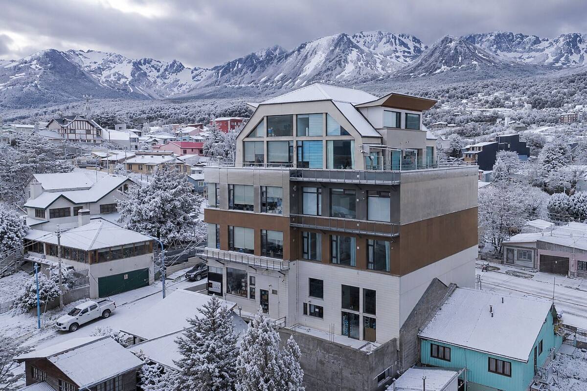 Un edificio de varios pisos con diseño moderno, situado en un pueblo nevado con montañas al fondo.