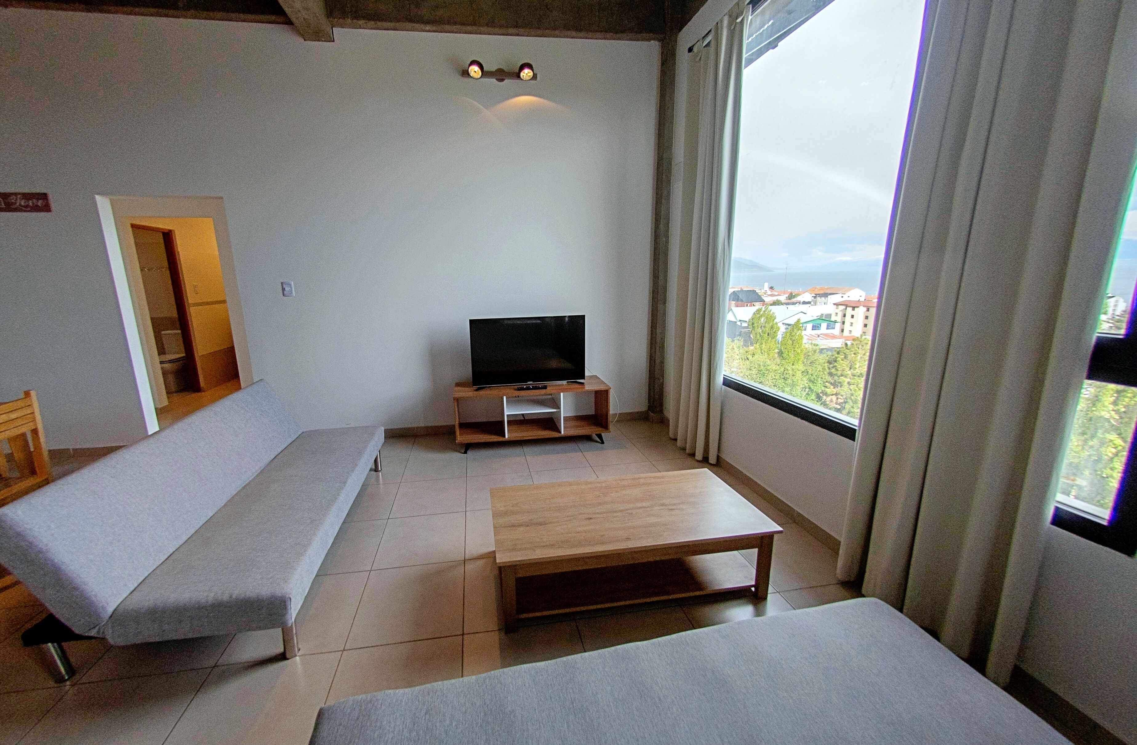 A modern living room with a sofa, a TV on a wooden stand, a coffee table, and a large window with curtains.