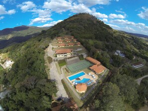 Vista aérea de un complejo turístico con varias construcciones, cancha de tenis y piscina rodeada de abundante vegetación.