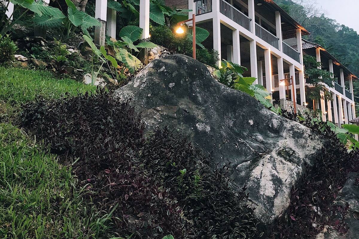 Un edificio de varios pisos con balcón, rodeado de exuberante vegetación y grandes rocas.
