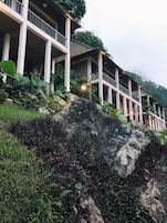 Um edifício com vários andares e varanda, rodeado por vegetação exuberante e pedras grandes.