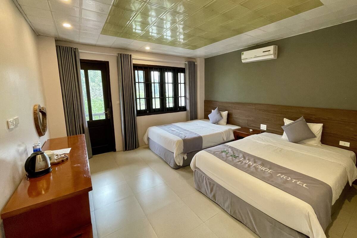 Habitación de hotel con dos camas, un escritorio, una silla, un ventanal con cortinas y un equipo de aire acondicionado.