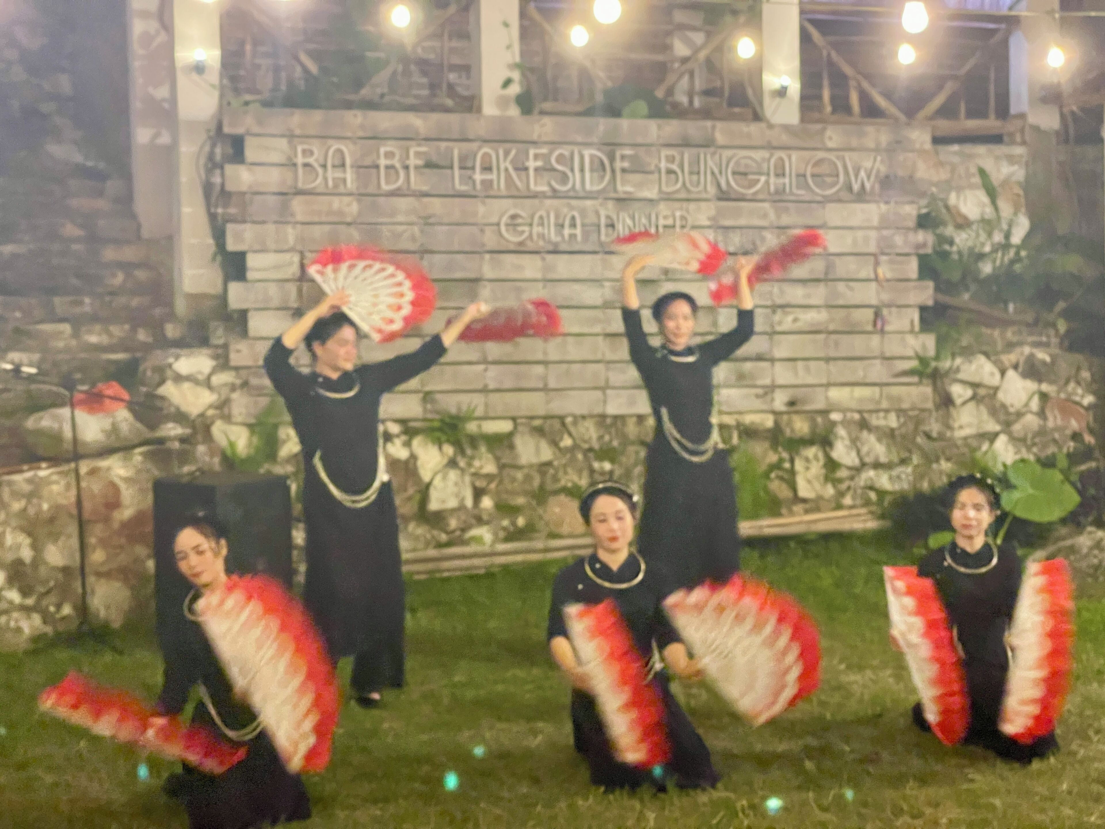 A group of people performing a dance with fans in front of a sign that reads "BA BE LAKESIDE BUNGALOW GALA DINNER".