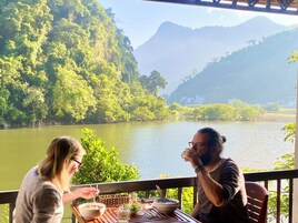 Uma área de refeições com mesa posta para dois, com vista para um rio e vegetação exuberante.