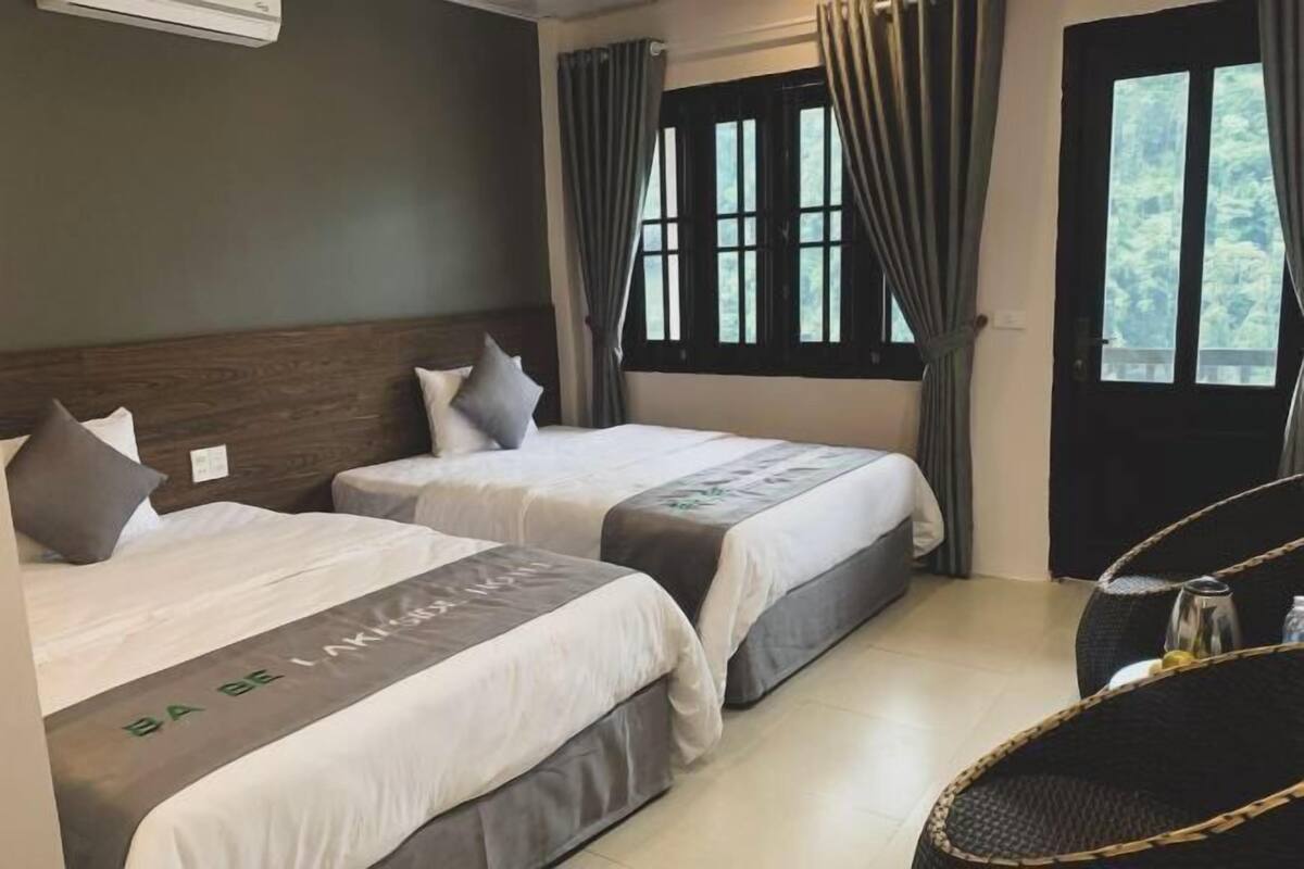 Habitación de hotel con dos camas, cabecero de madera, ventana con cortinas y una silla de mimbre.