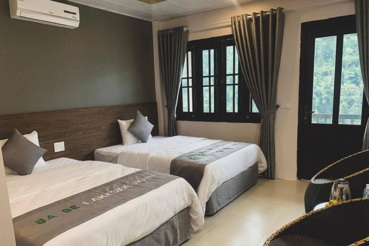 Una habitación de hotel con dos camas, una ventana con cortinas, una silla de mimbre y un aire acondicionado.