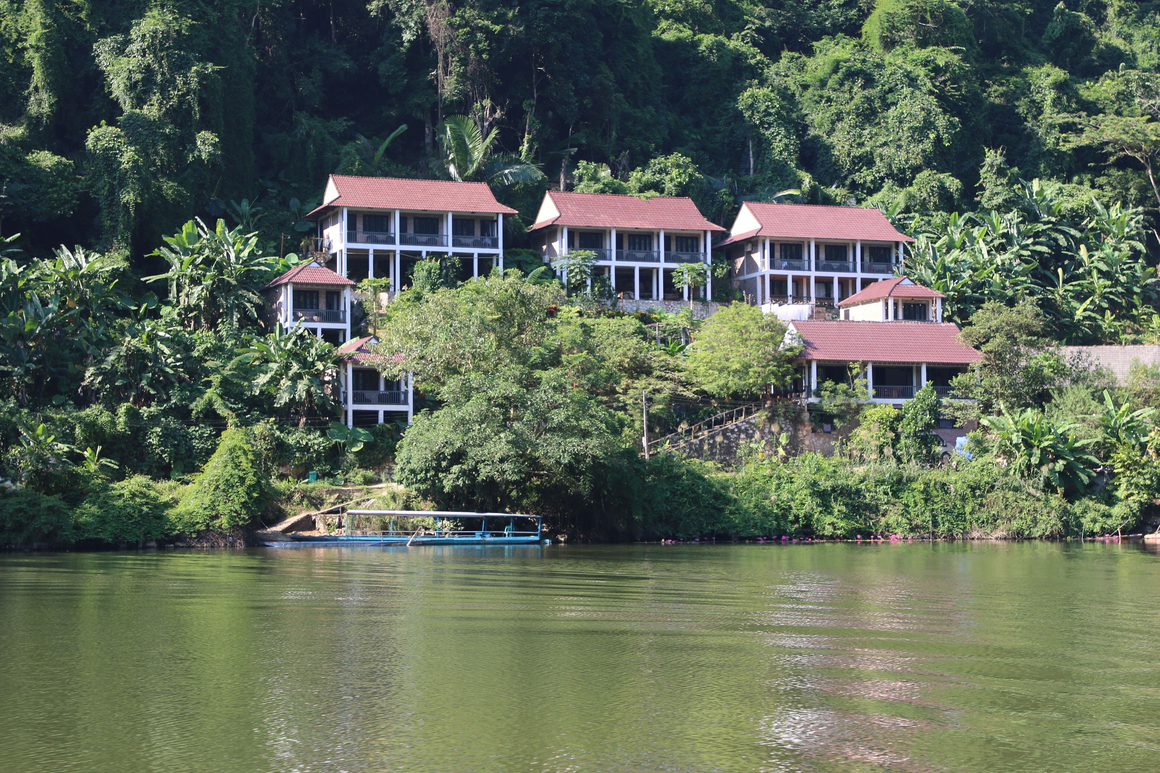 A row of white buildings with red roofs, surrounded by lush greenery and a body of water.