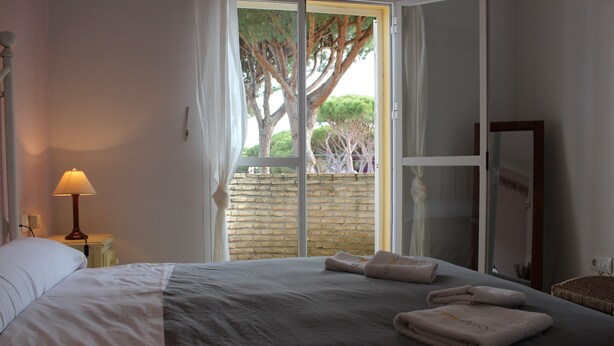 A bedroom with a large bed, a nightstand with a lamp, a window with a view of trees, and a mirror on the wall. - Spacious holiday home by the sea (Chiclana de la Frontera)
