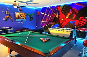 A pool table with a foosball table and a TV in a room with superhero-themed decorations.