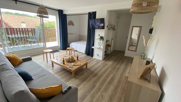 Un salon moderne avec un canapé, une table basse et un téléviseur. Il y a un balcon d’où l’on peut admirer un immeuble avec un toit rouge.