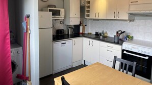 Une cuisine de taille réduite avec des meubles blancs, un four à micro-ondes, une cuisinière et un évier. Il y a une horloge au mur et un balai appuyé contre le mur.