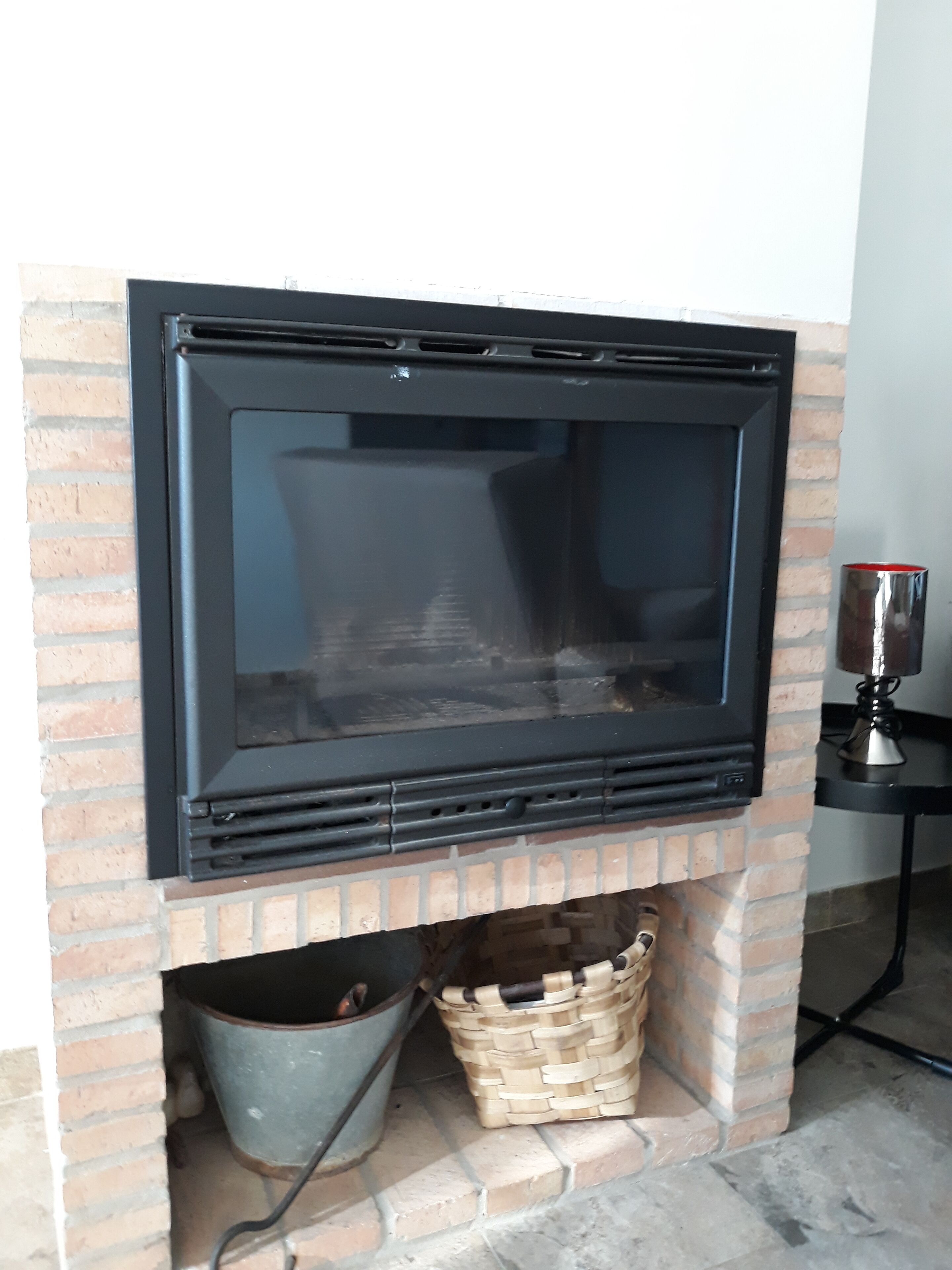 Une cheminée avec une façade en verre noir, un foyer en briques et, en dessous, un seau et un panier en métal.