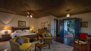 Quarto aconchegante com cama, sofá, mesa de centro e escrivaninha. Há uma porta que dá para uma varanda com área de estar externa.